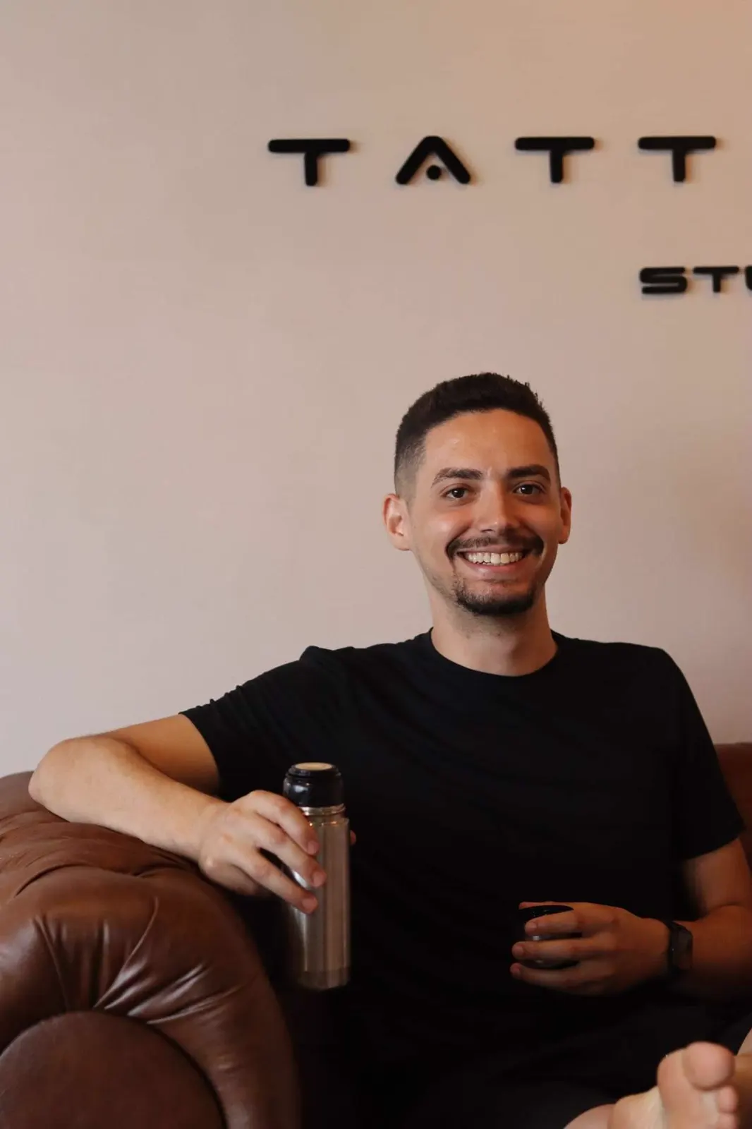 Matheus Borges sorrindo, sentado num sofá de estúdio com uma garrafa de café na mão.